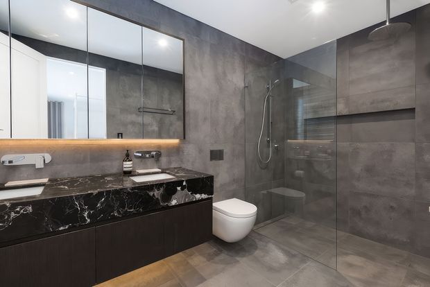 Contemporary bathroom with a floating dark timber vanity and dramatic black marble benchtop, paired with a wall-mounted basin tap and large frameless mirror. Large-format grey stone-look tiles cover the walls and floor, while a clear glass shower enclosure includes a handheld shower and recessed shelving. A wall-hung toilet and warm under-mirror LED lighting complete the refined, moody interior.