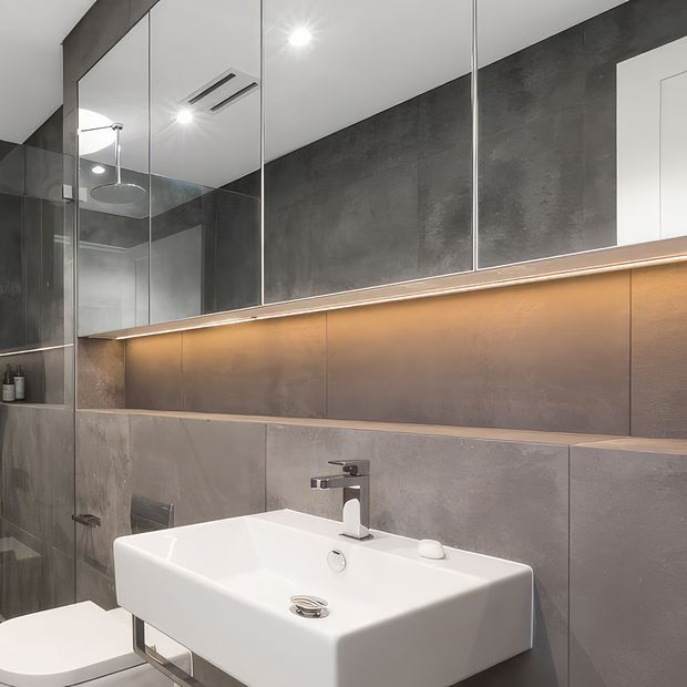 Contemporary bathroom with a wall-mounted white rectangular basin, chrome tapware, large grey concrete-look wall tiles, mirrored cabinetry, and warm integrated strip lighting beneath the mirror. A glass-enclosed shower is visible at left, with recessed ceiling lighting and a clean, minimalist fit-out.