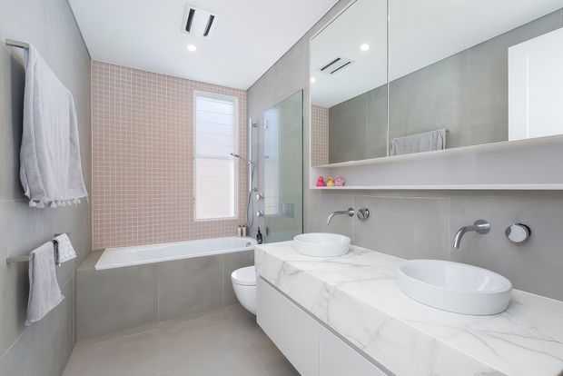 Bright contemporary bathroom with a double vanity, two vessel basins on a white marble-look benchtop, wall-mounted tapware, large mirrored cabinetry, a bathtub, shower enclosure and toilet. The room uses pale grey wall and floor tiles, a pink mosaic-tile feature wall, white cabinetry and recessed ceiling lighting.