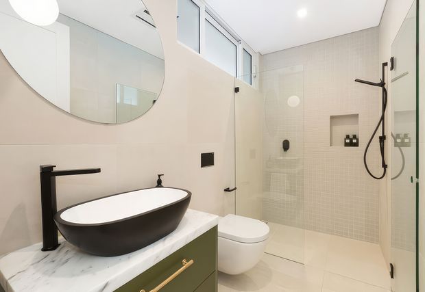 Contemporary bathroom featuring a freestanding black vessel basin on a white marble-look vanity, matte black tapware and shower fittings, a wall-hung toilet, pale micro-mosaic shower wall tiles, frameless glass shower screening, and a large rounded mirror. The palette is light and neutral with dark fixtures and subtle brass hardware accents.