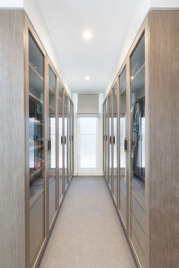 A bright, narrow walk-in wardrobe with symmetrical floor-to-ceiling glass-fronted storage cabinetry on both sides. The cabinets have vertically textured light timber finishes, slim dark frames, and brass-toned handles, with clothing and accessories visible inside. A soft grey carpet runs through the central aisle to a window with a roller blind. Recessed ceiling lights provide even illumination, creating a clean, refined and highly organised appearance.