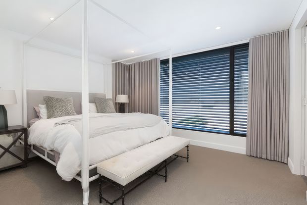 Bright, refined bedroom with a white upholstered bed, mirrored wardrobe doors, neutral carpet, bedside tables and lamps, full-height curtains, and large windows with horizontal blinds. The restrained palette and clean-lined furnishings create an airy, serene feel.