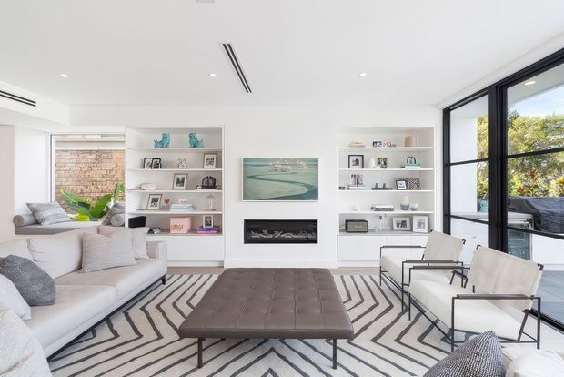 Bright, contemporary living room with a large white sectional sofa, two minimalist lounge chairs, a substantial tufted leather ottoman, and a bold black-and-white geometric rug. White built-in shelving flanks a central fireplace and artwork, while black-framed floor-to-ceiling glazing opens toward an outdoor balcony or terrace. The room has a clean, airy, refined feel with abundant natural light.