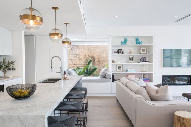 Bright open-plan contemporary interior combining a white kitchen with a large marble-look island, black bar stools, pendant lighting, and a neutral living area. Built-in shelving displays framed photographs and decorative objects, while large glazing opens to a planted courtyard or exterior view. A fireplace and wall-mounted artwork are visible in the living zone.