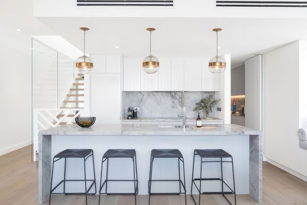 Bright contemporary open-plan kitchen with a large marble-look island, four black metal bar stools, white flat-panel cabinetry, and three brass pendant lights. A glass-sided staircase is visible to the left, with light timber flooring and a marble splashback behind the sink and cooktop.