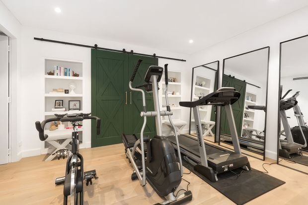 Bright, compact home gym with timber flooring, white walls, built-in open shelving, and a dark green barn-style sliding door. The room contains an exercise bike, elliptical trainer, treadmill, mirrors, and workout mats.