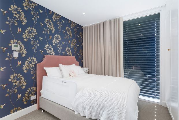 A styled bedroom with a white upholstered bed and blush pink curved headboard, set against a dramatic navy floral feature wall. Full-height beige curtains frame a large window with white horizontal blinds, while neutral carpet and white bedding create a bright, comfortable interior.