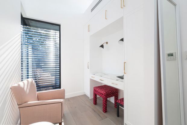 Bright compact study nook or home office with built-in white cabinetry, a white desk, brass hardware, integrated black wall lamps, and patterned red stools. A blush upholstered armchair sits beside a window with horizontal blinds, while pale timber-look flooring and white walls create an airy interior.