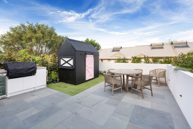 A bright rooftop-style outdoor terrace paved in large grey stone slabs, enclosed by white rendered walls. The space includes a timber dining table with woven chairs, a compact black gabled garden shed with a pink door, and an outdoor kitchen or barbecue unit at the edge. Mature greenery and neighbouring pitched-roof buildings are visible beyond the terrace.