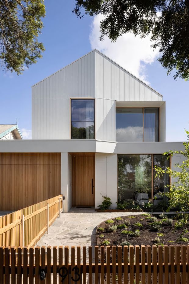 Front exterior view of a contemporary two-storey residence with a steep gabled roof clad in pale vertical panels, large dark-framed windows, warm timber garage and entry doors, and a landscaped front garden. A timber picket fence and pedestrian gate frame the foreground.
