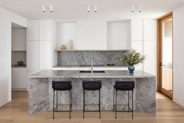 Bright, refined contemporary kitchen featuring a large grey stone island with waterfall ends, integrated sink and three black bar stools. White full-height cabinetry surrounds a matching stone splashback and open display shelf, with light timber flooring and a timber-framed glazed door.