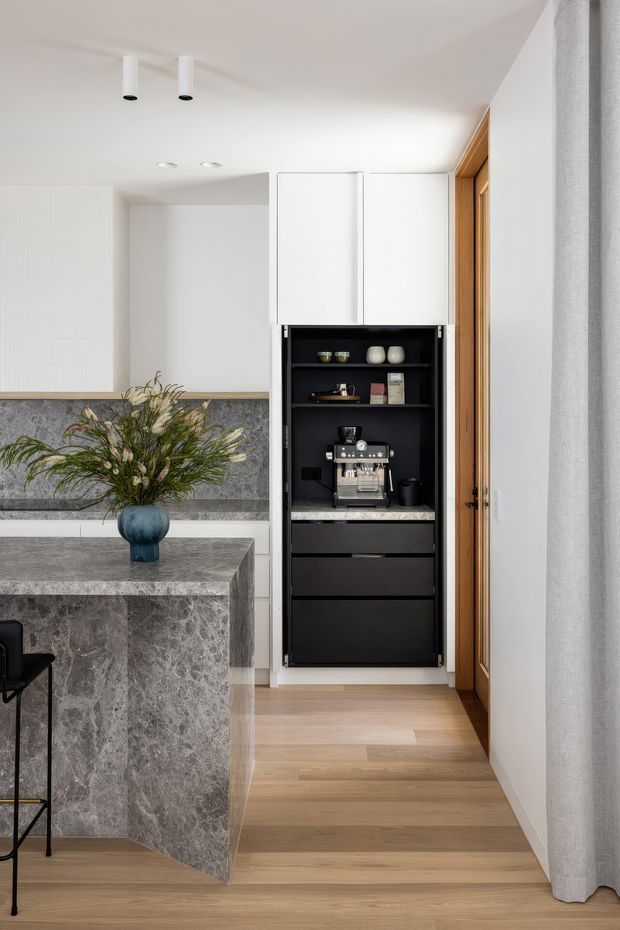 Bright contemporary kitchen interior featuring a large grey-veined stone island and matching backsplash, white upper cabinetry, a dark integrated coffee station, warm timber flooring, and a timber-framed doorway beside a full-height curtain. A blue vase with foliage provides a small decorative accent.