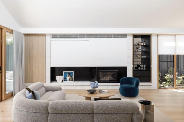 A bright contemporary living room with a large curved modular sofa arranged around a low timber coffee table, facing a built-in fireplace and television wall. The feature wall combines white panelled surfaces, light timber cabinetry, black open shelving and a recessed fireplace. Full-height glazing and sheer curtains bring in natural light, with a blue accent armchair and indoor planting adding colour.