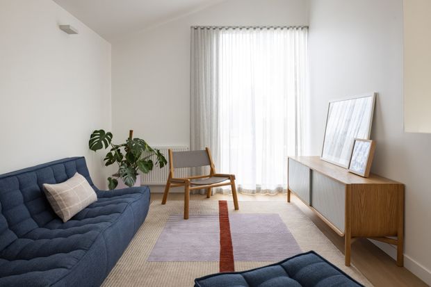 Bright, minimalist living room with a deep blue tufted sofa and matching ottoman, a pale patterned area rug, a wooden mid-century-style armchair, and a timber sideboard with framed artwork. Sheer floor-to-ceiling curtains diffuse abundant natural light, while indoor plants add a natural accent.