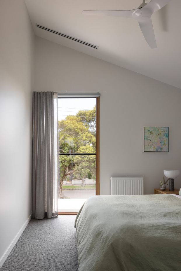 A bright, minimalist bedroom with a sloped white ceiling, ceiling fan, integrated linear air-conditioning vent, soft grey carpet, and a large glazed timber-framed door opening to greenery outside. The room contains a neatly made bed with a pale textured cover, grey curtains, a radiator, and a small bedside table with a lamp and artwork.