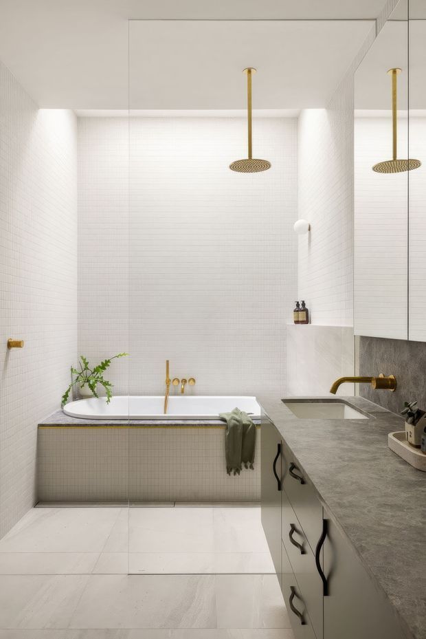 A bright, refined bathroom featuring a freestanding bathtub clad in small white tiles, full-height white tiled walls, large pale floor tiles, and a long grey stone vanity with integrated basin. Brushed brass shower hardware, taps, and fixtures provide warm accents, while a large mirror reflects the shower area. The room has a clean contemporary feel with subtle greenery and minimal accessories.