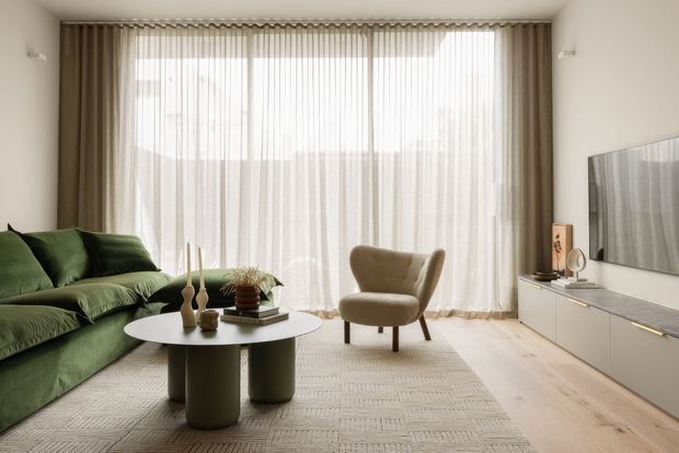 A bright, refined contemporary living room with a large wall of sheer floor-to-ceiling curtains, pale timber flooring, a textured neutral rug, a deep green sofa, sculptural round coffee tables, and a beige upholstered accent chair. A wall-mounted television and low built-in media cabinetry occupy the right wall.