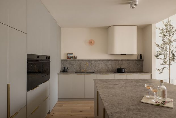 A refined contemporary kitchen with full-height matte cabinetry, integrated black oven, stone benchtops and splashback, brass-toned tapware and handles, and a large central island. Warm timber flooring and soft neutral finishes create a calm, minimalist space, with abundant daylight entering from the glazed opening at right.