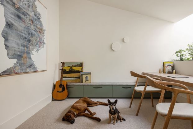 Bright, minimalist dining and living area with a long built-in muted green storage cabinet, pale stone or composite countertop, timber dining table and Scandinavian-style wooden chairs. A guitar, framed artwork, indoor plant, and two dogs add lived-in character. The room has white walls, a light carpeted floor, and soft natural light.