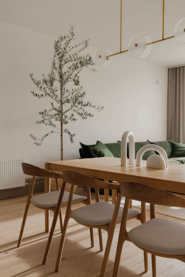 A warm, refined dining and living area with a long light-wood dining table surrounded by upholstered timber dining chairs. A dark green sofa sits behind the table against a white wall, accompanied by a tall indoor branch arrangement. The room features a sculptural linear pendant light with frosted circular shades, pale timber flooring, beige curtains, and minimalist ceramic or stone decorative forms on the table.
