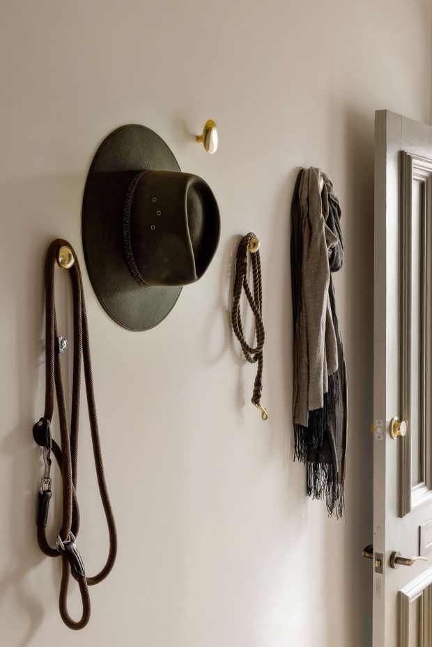 A close-up of a warm, traditional entryway wall beside a partially open paneled door. The neutral painted wall features decorative brass hooks holding a dark leather hat, a scarf, and equestrian-style leather tack or reins. The scene has refined vintage detailing, soft natural light, and a compact, welcoming feel.