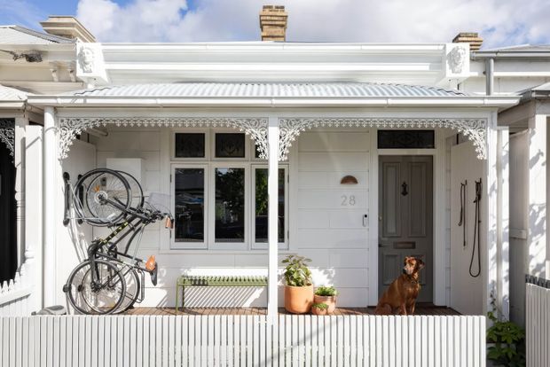 Bright street-facing facade of a narrow heritage terrace or bungalow-style residence, featuring white weatherboard cladding, ornate lacework verandah detailing, a corrugated metal roof, decorative brackets, timber-framed windows and a recessed grey front door. Bicycles, potted plants and a dog occupy the front verandah, with the house number 28 visible beside the entry.