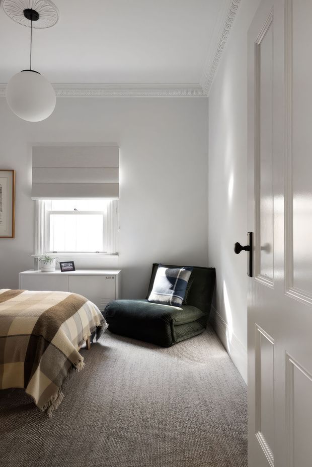 Bright, compact bedroom viewed from the doorway, featuring white walls and detailed decorative cornice moulding, a white paneled door, grey carpet, a small window with a Roman blind, a low white cabinet, a dark upholstered lounge chair, and a bed partially visible in the foreground with a neutral checkered throw. A simple round pendant light hangs from the ceiling.