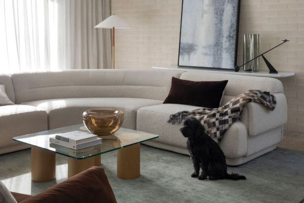 A bright, refined living room featuring a large cream boucle sectional sofa, glass coffee table with timber pedestal legs, neutral rug, sculptural floor lamp, abstract artwork, and a black dog. A pale painted brick feature wall and sheer curtains create a soft contemporary setting, with dark brown textiles adding contrast.