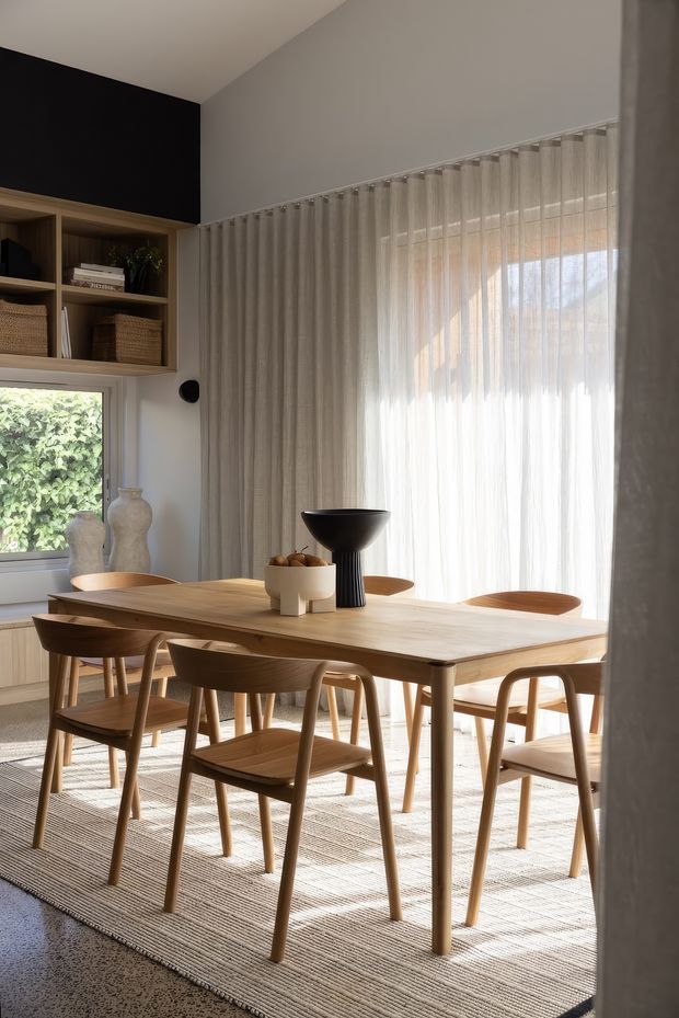 A warm, contemporary dining room with a large light timber dining table surrounded by matching wooden chairs. Sheer full-height curtains filter abundant daylight from a large window, while built-in timber shelving with woven baskets and decorative ceramics occupies the left wall. The room has pale walls, a dark charcoal feature wall, a textured neutral rug, and a black sculptural centerpiece on the table.