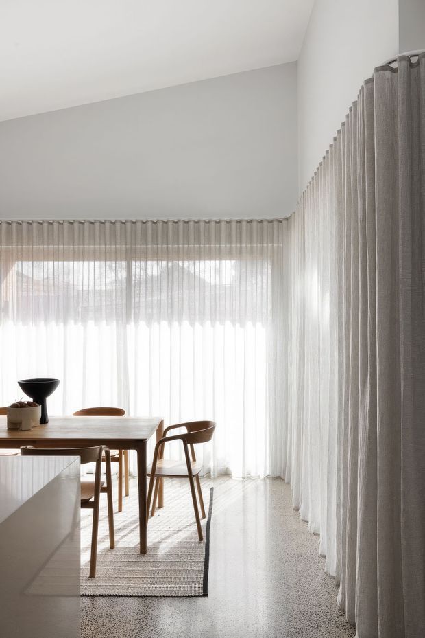 Bright minimalist dining area with a timber dining table and chairs positioned beside floor-to-ceiling sheer curtains. The room has white walls, abundant filtered daylight, a pale textured rug, and a polished light terrazzo or concrete floor. A white kitchen island or counter is partially visible in the foreground.