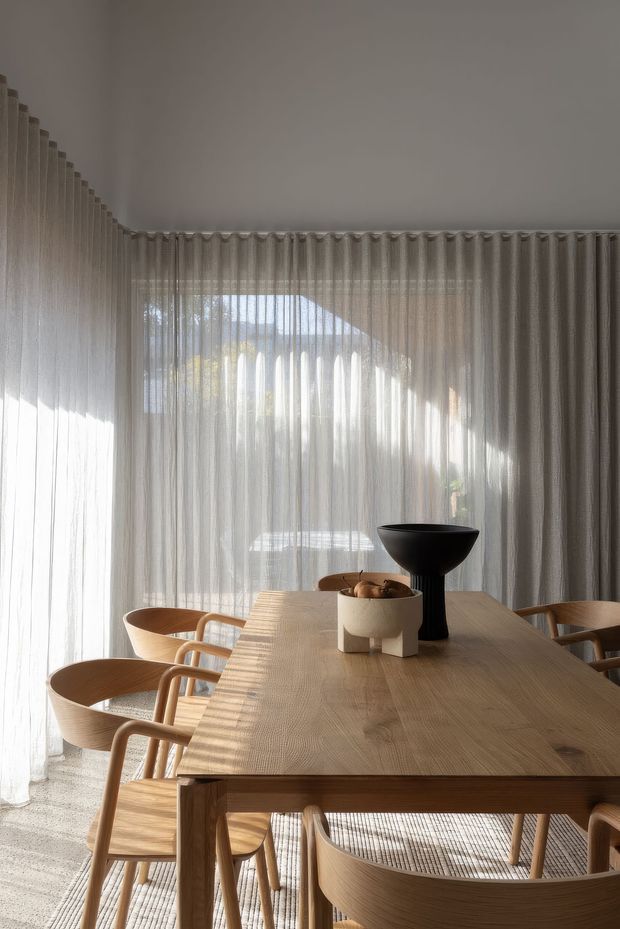 A warm, light-filled dining room with a large natural timber dining table surrounded by curved timber dining chairs. Full-height sheer curtains cover the glazing on two sides, filtering strong sunlight and creating soft shadows. Minimal sculptural black and neutral-toned objects sit on the table, while a pale textured rug covers the floor.
