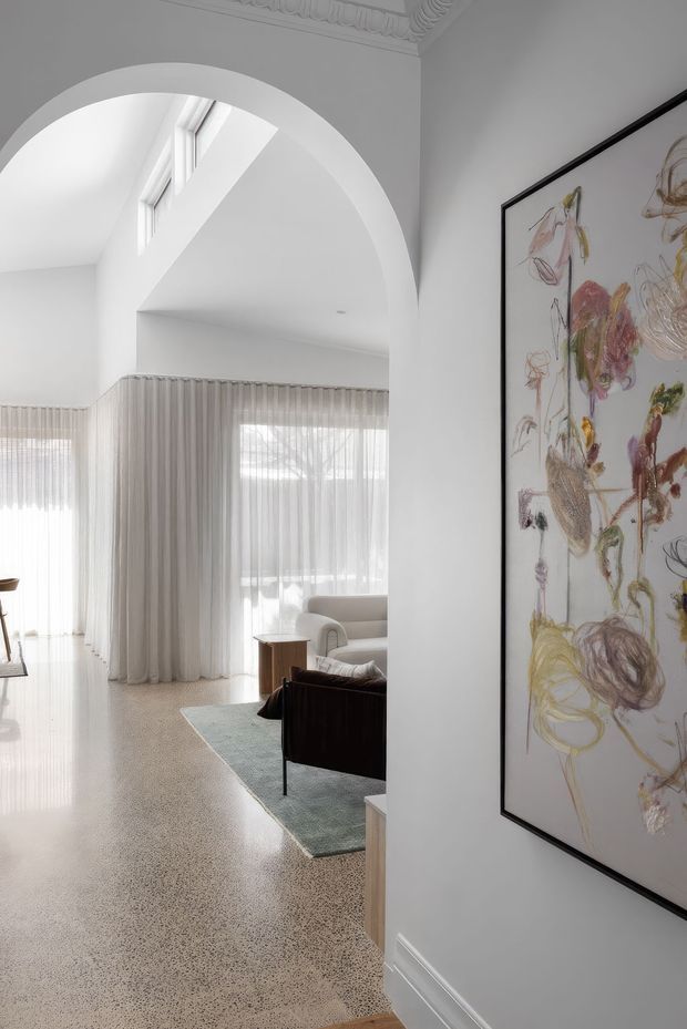 Bright, refined interior view through a large rounded archway into a living room. The space features white walls, ornate ceiling cornice detailing, full-height sheer curtains, polished light terrazzo flooring, a pale rug, sculptural neutral seating, dark wood furniture, and a large framed abstract artwork in the foreground. Tall glazing and abundant daylight create an airy, elegant atmosphere.