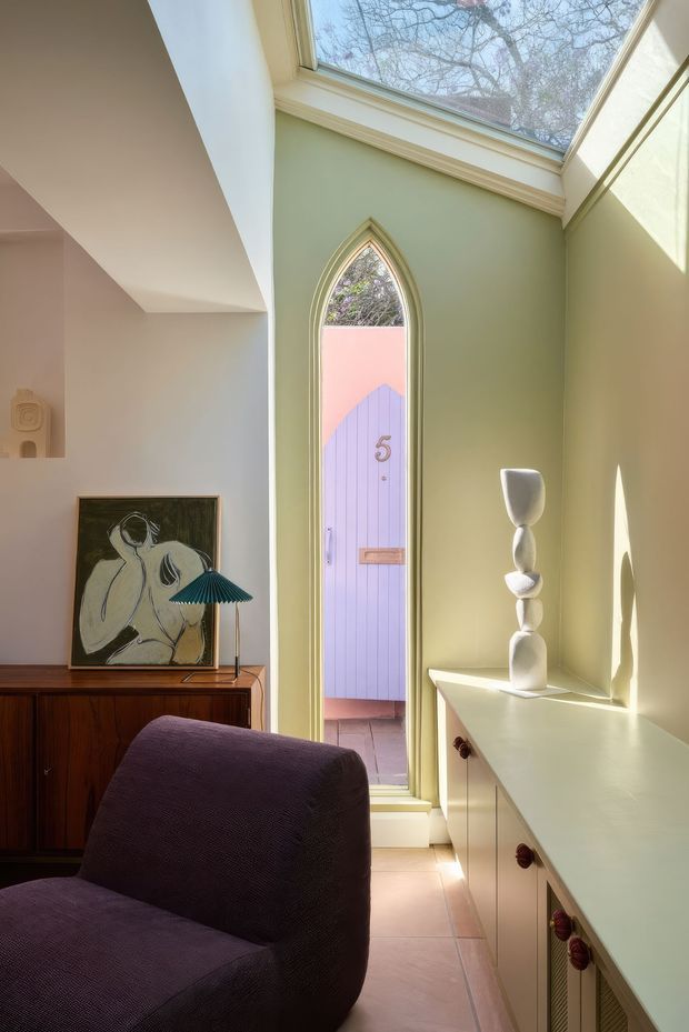 A warm, eclectic sitting area with a deep purple upholstered lounge chair, dark timber sideboard, framed artwork, and a small sculptural lamp. Pale sage-green walls surround a tall pointed-arch glazed door opening to an exterior courtyard, while a large overhead skylight brings in abundant natural light. A pale built-in cabinet runs along the right wall, topped with a sculptural white vase.