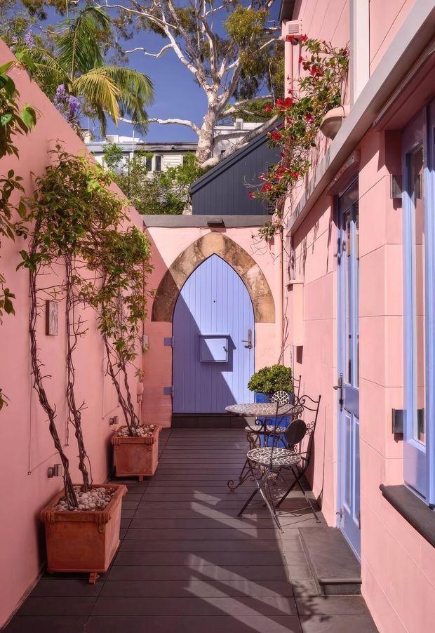 A narrow, sunlit courtyard passage finished in pink, with dark timber decking, climbing plants in terracotta planters, a small wrought-iron café table and chairs, and a lavender-blue arched door framed by a stone arch. The passage is enclosed by pink rendered walls and opens visually toward lush tropical planting and neighbouring structures under a clear blue sky.