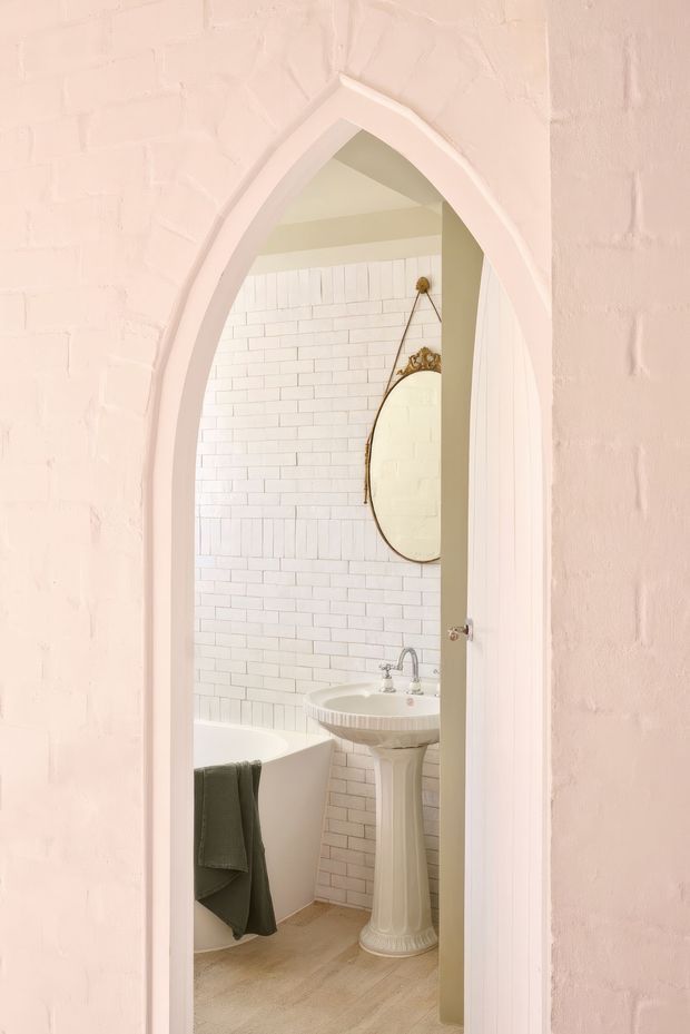 A soft, light-toned bathroom viewed through a pointed Gothic-style arched doorway. The room features white brick-pattern wall tiling, a freestanding pedestal basin with chrome tapware, a partially visible white bathtub, a decorative oval mirror, and pale timber-look flooring. The surrounding walls are painted blush pink with textured brickwork.