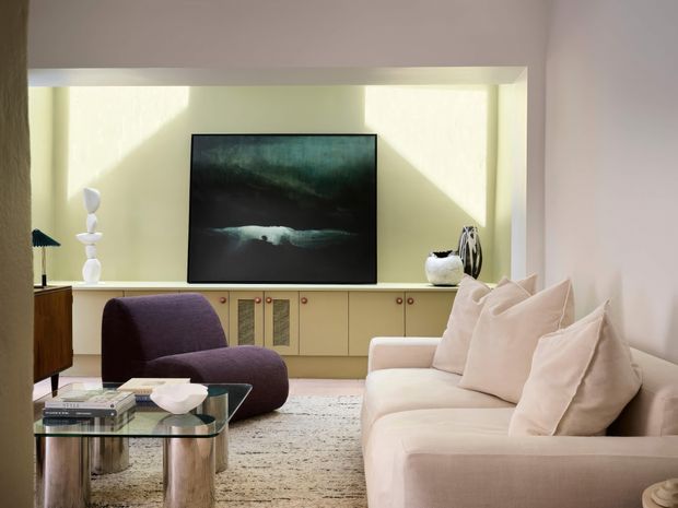A refined living room with a cream upholstered sofa and sculptural armchair arranged around a glass coffee table. A low, pale wood built-in media cabinet spans the rear wall beneath a dark abstract artwork, with soft daylight, neutral finishes, and contemporary decorative objects creating a calm, elegant atmosphere.