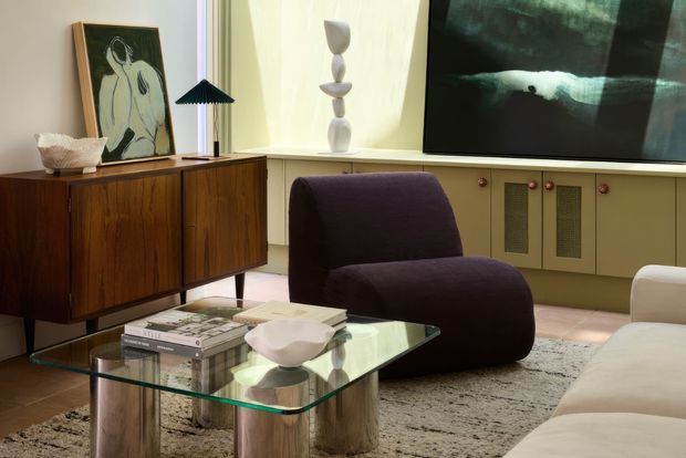 A refined living room featuring a mid-century-inspired timber sideboard, sculptural lounge chair, cream sofa, glass coffee table with cylindrical timber legs, textured rug, wall-mounted artwork, and built-in low cabinetry beneath a large artwork or screen. Warm natural light and earthy neutral tones create an inviting, curated atmosphere.
