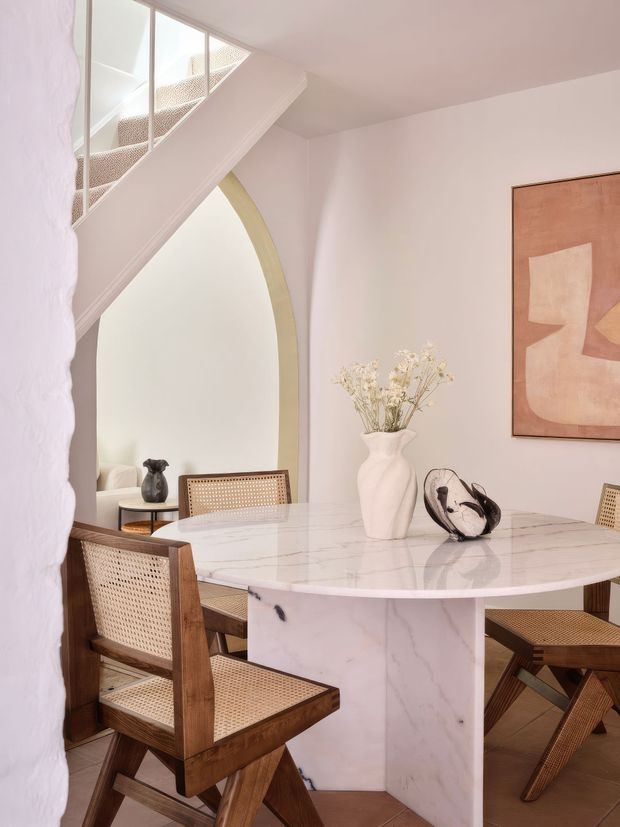 A bright, softly styled dining area with a large round marble-look pedestal table surrounded by warm timber and cane dining chairs. White walls, a sculptural arched opening, mirrored stair balustrade, abstract artwork, and restrained decorative objects create an airy, refined interior.