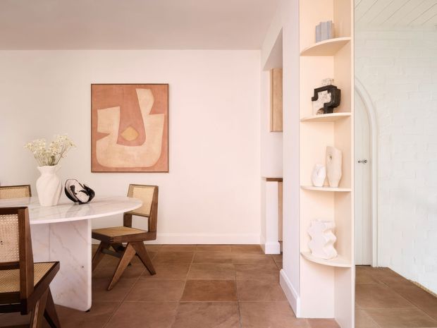 Warm, softly lit interior showing a dining area with a white marble-look table and woven leather-and-wood dining chairs. A large abstract artwork hangs on a pale wall, while a built-in curved display shelving unit holds sculptural decor and separates the dining space from an adjoining white-brick-lined area. The floor is finished in large brown tiles, and the overall scheme is refined and contemporary with organic, sculptural details.