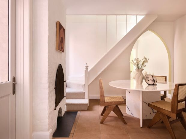 Bright, refined dining and stair area with a sculptural white dining table, woven-seat timber chairs, tiled flooring, a curved arched opening, and a white-painted staircase with glazed balustrade panels. The space has soft warm neutrals, textured white walls, and restrained contemporary detailing.