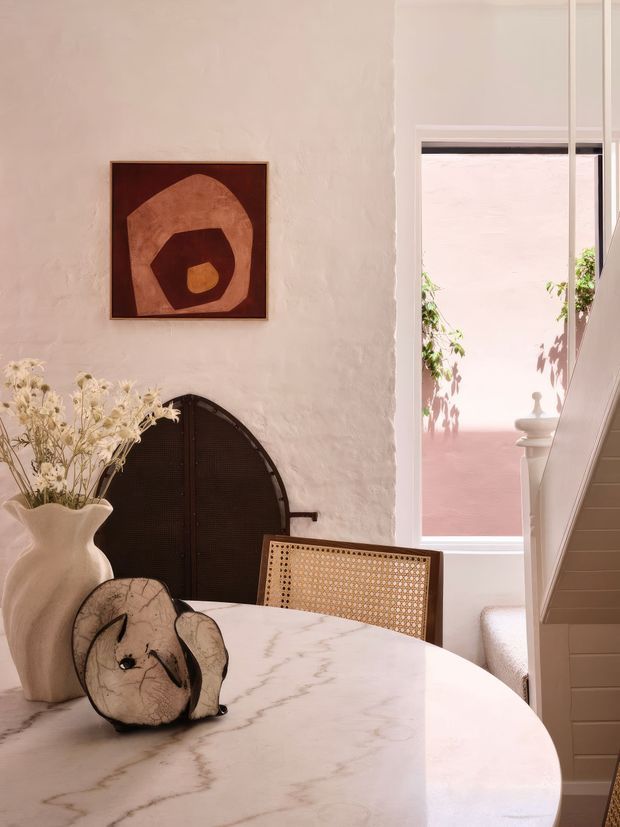 A softly lit dining area with a round pale stone dining table, woven cane dining chair, sculptural ceramic vase, dried flowers, and abstract artwork. The room features textured plaster walls, a dark arched built-in door or cabinet, and a stairway beside a large window overlooking a pink-toned courtyard. The overall aesthetic is warm, refined, and contemporary with natural materials and organic forms.