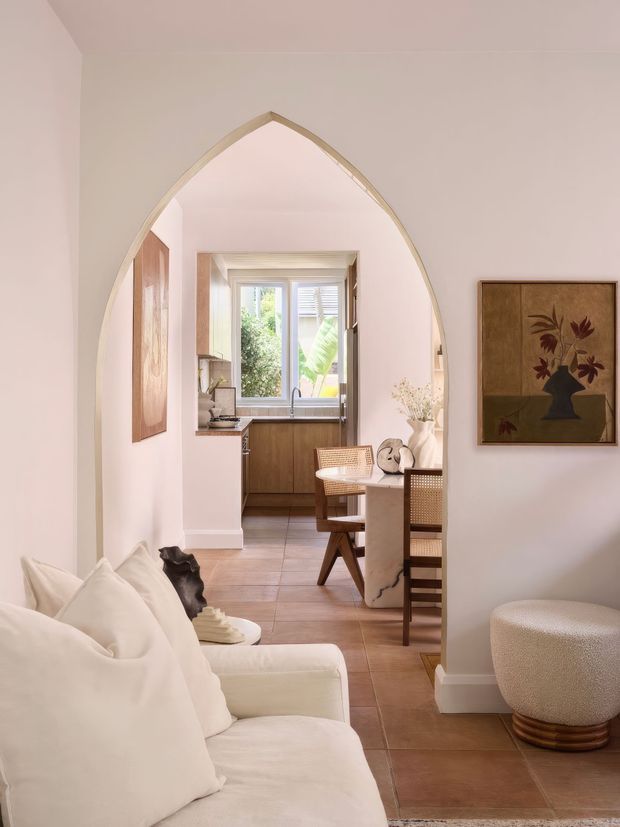 A softly styled interior view through a tall pointed archway, showing a light living room in the foreground, a dining area beyond, and a compact kitchen at the rear. The room features pale plaster walls, a warm tiled floor, cream upholstery, a marble-topped dining table, timber cabinetry, cane dining chairs, framed botanical artwork, and an arched architectural opening.