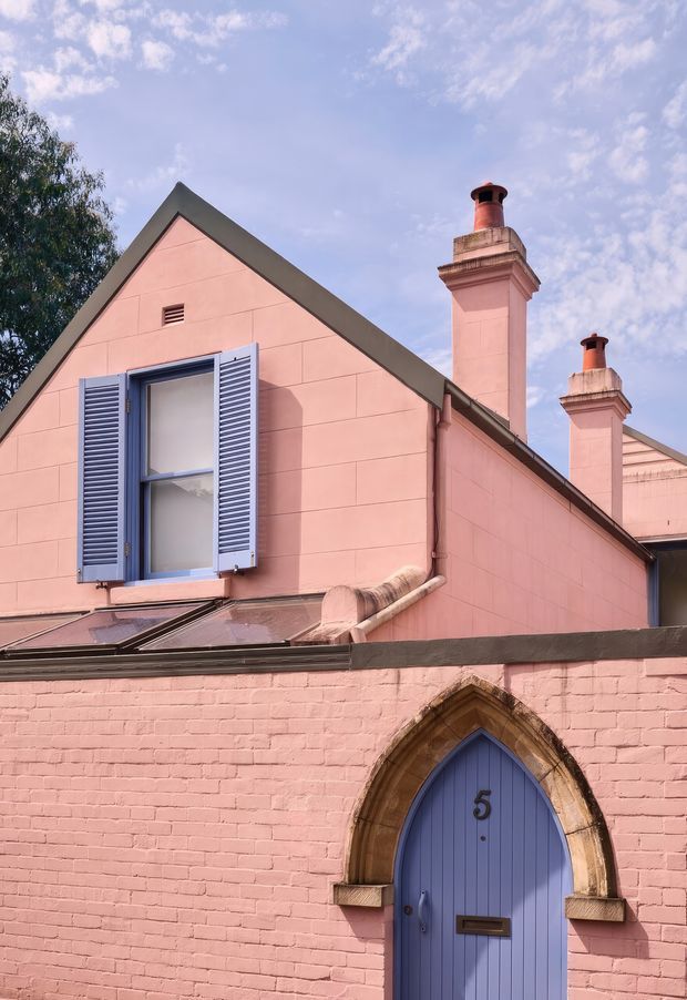 Exterior view of a distinctive pink rendered and brick building with steep gabled roof sections, ornate chimney stacks, blue louvred shutters, and a pointed Gothic-arched blue entrance door set within a stone surround.
