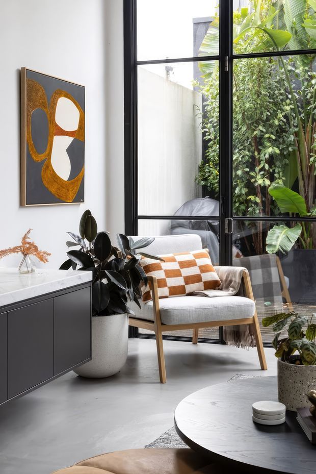 A bright, refined living room with a light grey upholstered timber armchair, patterned rust-and-cream cushion, dark built-in sideboard, round dark coffee table, indoor plants, and abstract artwork. Large black-framed steel-look glazing opens visually onto a lush courtyard garden. The room combines polished concrete flooring, white walls, and warm wood and ochre accents.