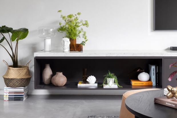 Contemporary living room vignette featuring a long dark built-in media console with open display shelving, a pale stone or marble-look benchtop, and curated decor. A wall-mounted television is partially visible above, with a round dark coffee table, upholstered seating, indoor plants, books, and decorative ceramics. The palette is monochromatic with warm natural accents.