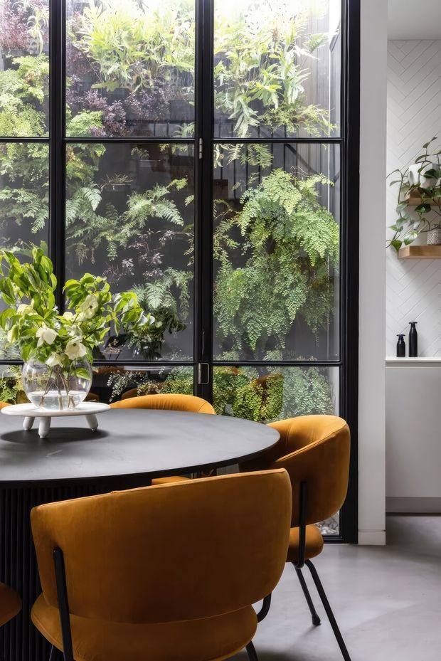 A stylish dining area with a round dark table and mustard velvet dining chairs positioned beside tall black-framed glazed doors or windows. Lush greenery is visible outside, while the interior includes white herringbone wall cladding, a floating wood shelf with potted plants, and a minimalist grey floor. The space has a refined, contemporary feel with strong indoor–outdoor connection and abundant natural light.
