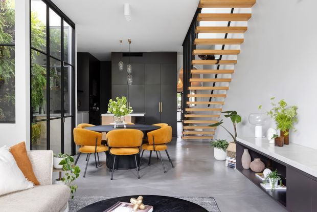 Bright contemporary open-plan interior combining a living area, dining area and kitchen. The space features a floating timber staircase with black metal supports, floor-to-ceiling black-framed glazing, polished grey concrete flooring, dark minimalist kitchen cabinetry, a mustard-gold upholstered dining set, and a sleek low media console with decorative objects and plants.