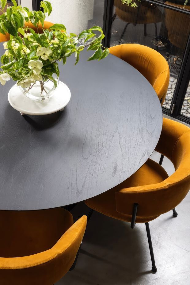 A close-up dining area with a large round dark table surrounded by mustard-orange upholstered dining chairs on slim black legs. A white ceramic vase filled with leafy green and white-flowering branches sits on the tabletop. The space is beside black-framed glazing, with a pebble-filled exterior lightwell or courtyard visible beyond. Strong natural light creates a warm, refined atmosphere.