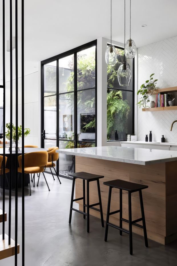 Bright contemporary kitchen with a large white island, pale timber cabinetry, black bar stools, black-framed floor-to-ceiling glazing, and a white herringbone-tile backsplash. A small dining area with mustard upholstered chairs is visible beyond a black vertical partition, with pendant lights and greenery adding warmth.