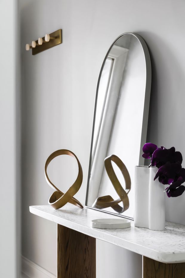 Refined entryway vignette featuring a slim white stone console with dark timber supports, a tall arched mirror, sculptural brass decor, a ribbed white vase with deep purple orchids, and a small brass wall light. The setting is bright, minimalist, and softly contemporary.